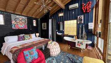 A bedroom with a bed, a red bean bag, a large window, a bathtub, and a vanity with a mirror.
