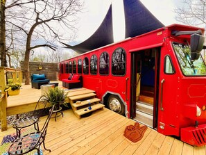 A red bus converted into a stationary living space with a deck, seating area, and a small garden.