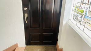 Una puerta de madera oscura con manija metálica y cerradura, un felpudo pequeño en la entrada y un ventanal con barrotes que muestra vista a la calle.