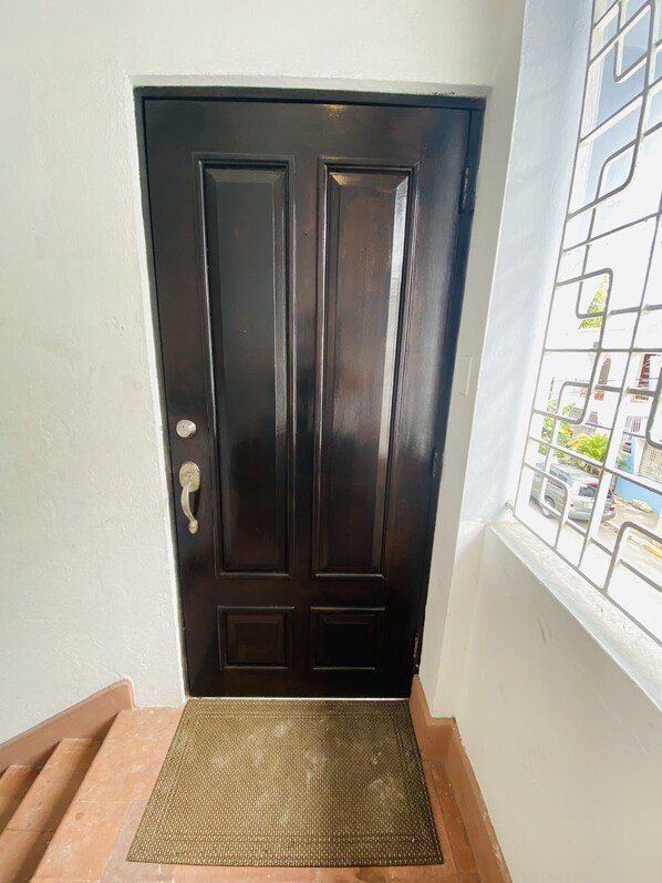 A dark wooden door with a metal handle and a keyhole, a small mat at the entrance, and a window with metal bars showing a street view. - Fiesta Spot at La Placita- Remodeled and convenient located spot. (San Juan)