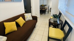 Una sala de estar con un sofá marrón, cojines amarillos, una pequeña mesa redonda con una silla y una zona de comedor al fondo.