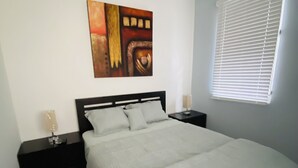 Un dormitorio con cama, mesita de noche, un cuadro en la pared y una ventana con persianas.