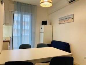 Ein kleiner, moderner Raum mit einem weißen Tisch, zwei Stühlen, einem blauen Sofa, einem Kühlschrank, einem Bild mit Strandmotiv und einem Fenster mit durchsichtigen Vorhängen.