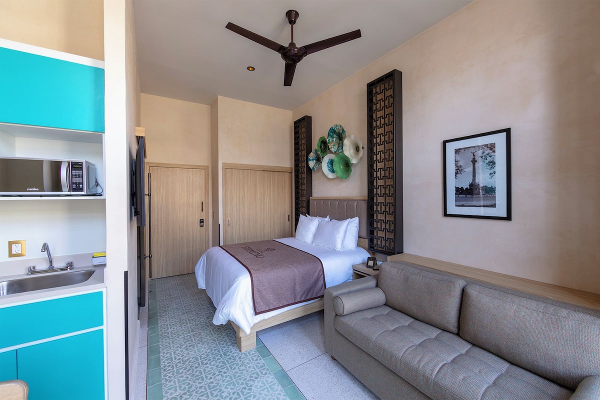 A hotel room with a bed, a sofa, a microwave, a sink, and a ceiling fan.