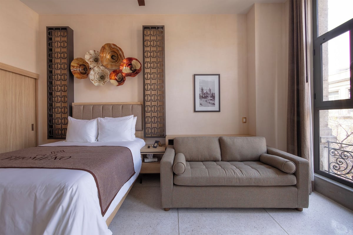 A hotel room with a bed, a sofa, a nightstand, decorative wall panels, and a framed picture.