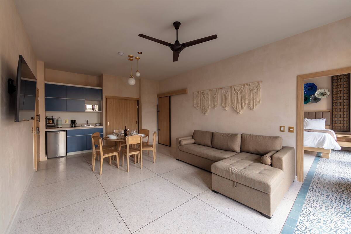 A modern living room with a sofa, dining table, and chairs. There is a kitchen area with cabinets and a microwave. A ceiling fan and pendant lights are visible.