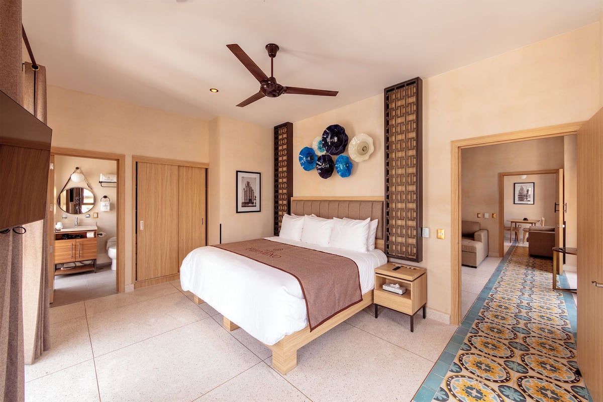 A hotel room with a bed, bedside tables, a ceiling fan, a bathroom with a toilet and sink, and a hallway with a patterned floor.