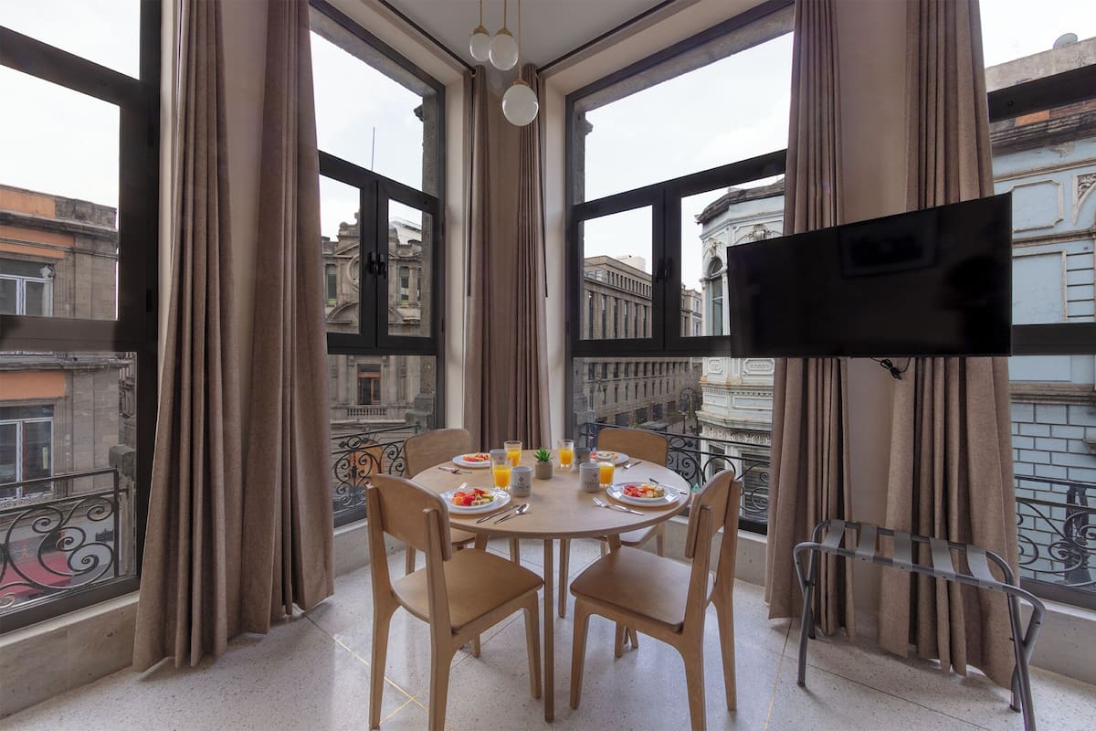 A dining area with a table set for four, a TV mounted on the wall, and a view of the city through large windows.