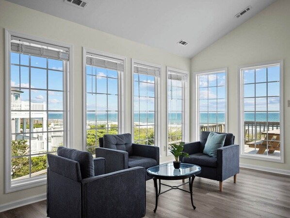 A spacious room with large windows offering ocean views, a seating area with sofas and a coffee table, and a wooden floor.