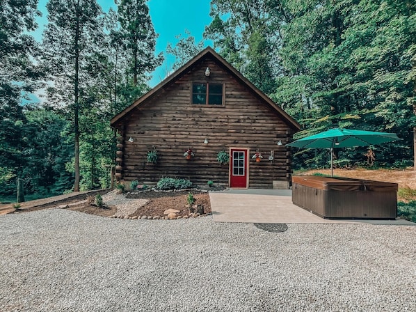 Une cabane en rondins avec une porte rouge, entourée d’une allée gravillonnée et d’un petit jardin.