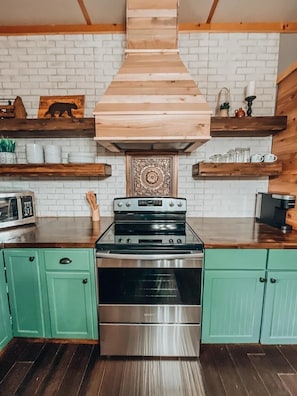Une cuisine dotée d’une cuisinière en acier inoxydable, de meubles de rangement verts, d’étagères en bois et d’un carrelage décoratif mural.