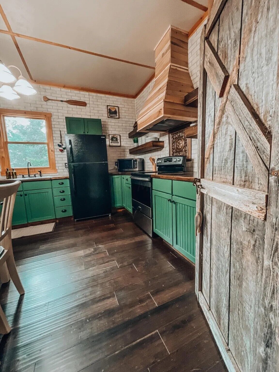 Eine rustikale Küche mit grünen Schränken, einem schwarzen Kühlschrank, Holzregalen und einer großen Holztür.