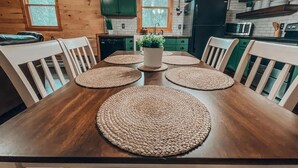 A wooden dining table with woven placemats, surrounded by white chairs, in a kitchen with green cabinets and a black refrigerator. - Rustic Oak Cabin - dogs welcome - Hocking hills (Logan)