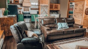 A cozy living room with a leather sofa, a dining table, and a kitchen in the background. - Rustic Oak Cabin - dogs welcome - Hocking hills (Logan)