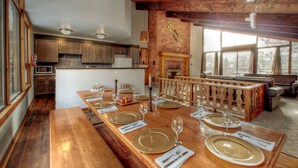A spacious dining area with a long wooden table set for a meal, a fireplace, and a view of the outdoors through large windows. - Spacious 8 bdrm Cottage with Private Beach Access, BBQ, Hot Tub, Sauna, Pet friendly (The Blue Mountains)