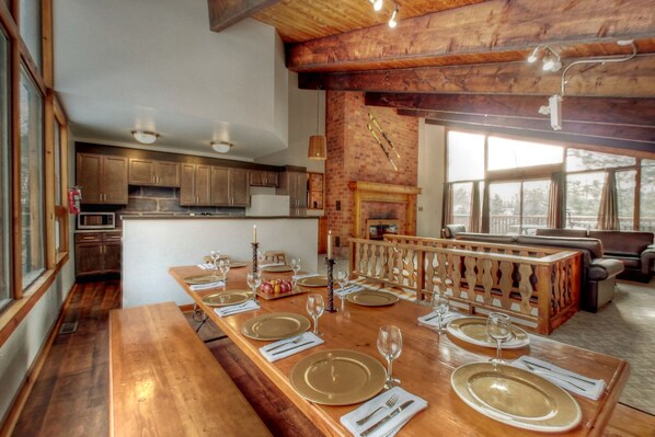 A spacious dining area with a long wooden table set for a meal, a fireplace, and a view of the outdoors through large windows. - Spacious 8 bdrm Cottage with Private Beach Access, BBQ, Hot Tub, Sauna, Pet friendly (The Blue Mountains)