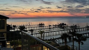Blick auf das Wasser mit einer Mole, die sich ins Wasser erstreckt, Palmen und einem Gebäude mit einem Balkon.
