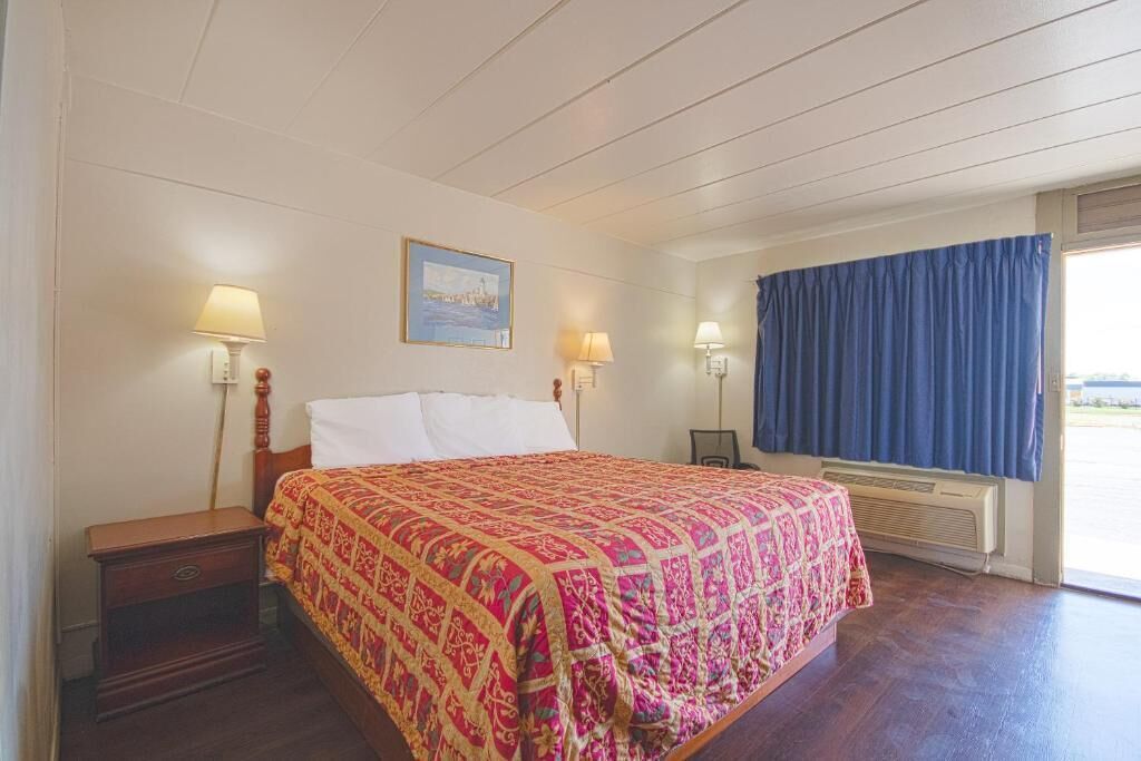 A hotel room with a bed, two bedside lamps, a window with curtains, and a painting on the wall.