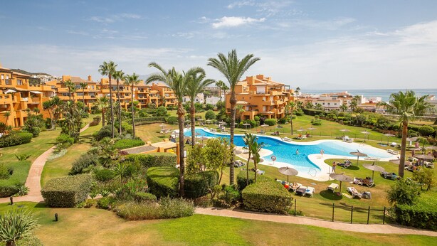 A resort with a central swimming pool surrounded by palm trees and sun loungers.
