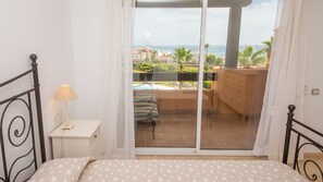 Une chambre avec un lit, une table de chevet, une lampe et une vue sur une piscine et l’océan, visible à travers une porte-fenêtre coulissante.