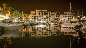A marina with boats docked, surrounded by multi-story buildings with balconies and a calm water surface reflecting the structures.