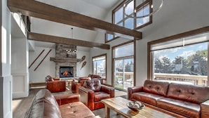 A spacious living room with leather sofas, a fireplace, large windows, and a high ceiling with exposed beams. - Brand New 7 bdrm Luxury Cottage with Private Beach Access, BBQ,7.5 baths, Hot Tub, Ping Pong Table (The Blue Mountains)