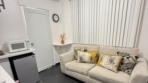 Un espace de vie compact comprenant un coin cuisine, un canapé agrémenté de coussins à motifs, un petit tapis et une fenêtre avec des stores.