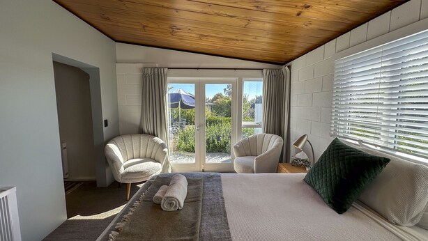 A bedroom with a large bed, two armchairs, a wooden ceiling, and a view of the outdoors through a glass door. - Sunset Sanctuary - Havelock North Holiday Home (Havelock North)