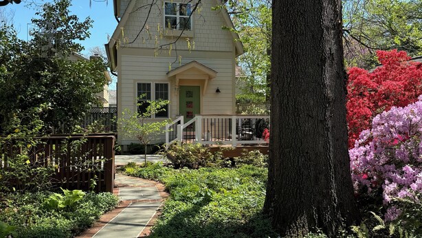A two-story house with a front porch, surrounded by lush greenery and flowering trees.