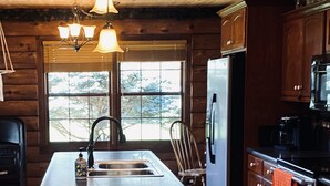 A kitchen with a central island, stainless steel appliances, wooden cabinetry, and a large window with a view of trees. - Log Cabin Haus, Amish Country. 
Shipshewana, Indiana near Pumpkinvine Trail (Shipshewana)