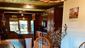 A wooden dining table with chairs, a vase with a plant, and a kitchen in the background. - Log Cabin Haus, Amish Country. 
Shipshewana, Indiana near Pumpkinvine Trail (Shipshewana)