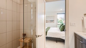 Une salle de bain moderne avec une douche à parois de verre, un tabouret en bois et un lit avec des coussins verts dans la chambre adjacente.
