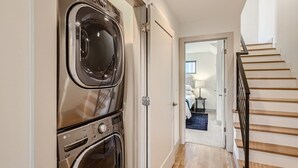 Une buanderie équipée de deux machines à laver à chargement frontal, un couloir menant à une chambre avec un lit, une table de chevet et une lampe.