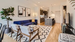 Un salon moderne avec un canapé bleu, un coin repas avec une table et des chaises, une cuisine en arrière-plan et un escalier menant à un autre niveau.