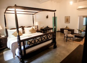A four-poster bed with a wooden frame, a bedside table with a lamp, a chair, and a desk with a vase of flowers. - Kathaluwa Grand Manor (Koggala)