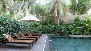 Zona junto a la piscina con sillones de madera y una sombrilla grande, rodeada de exuberante vegetación y palmeras.