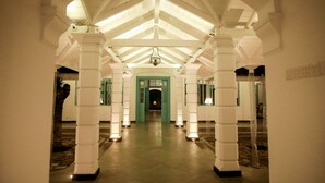 Un pasillo con columnas blancas y una puerta verde al final, que conduce a una habitación con techo alto y una lámpara colgante.
