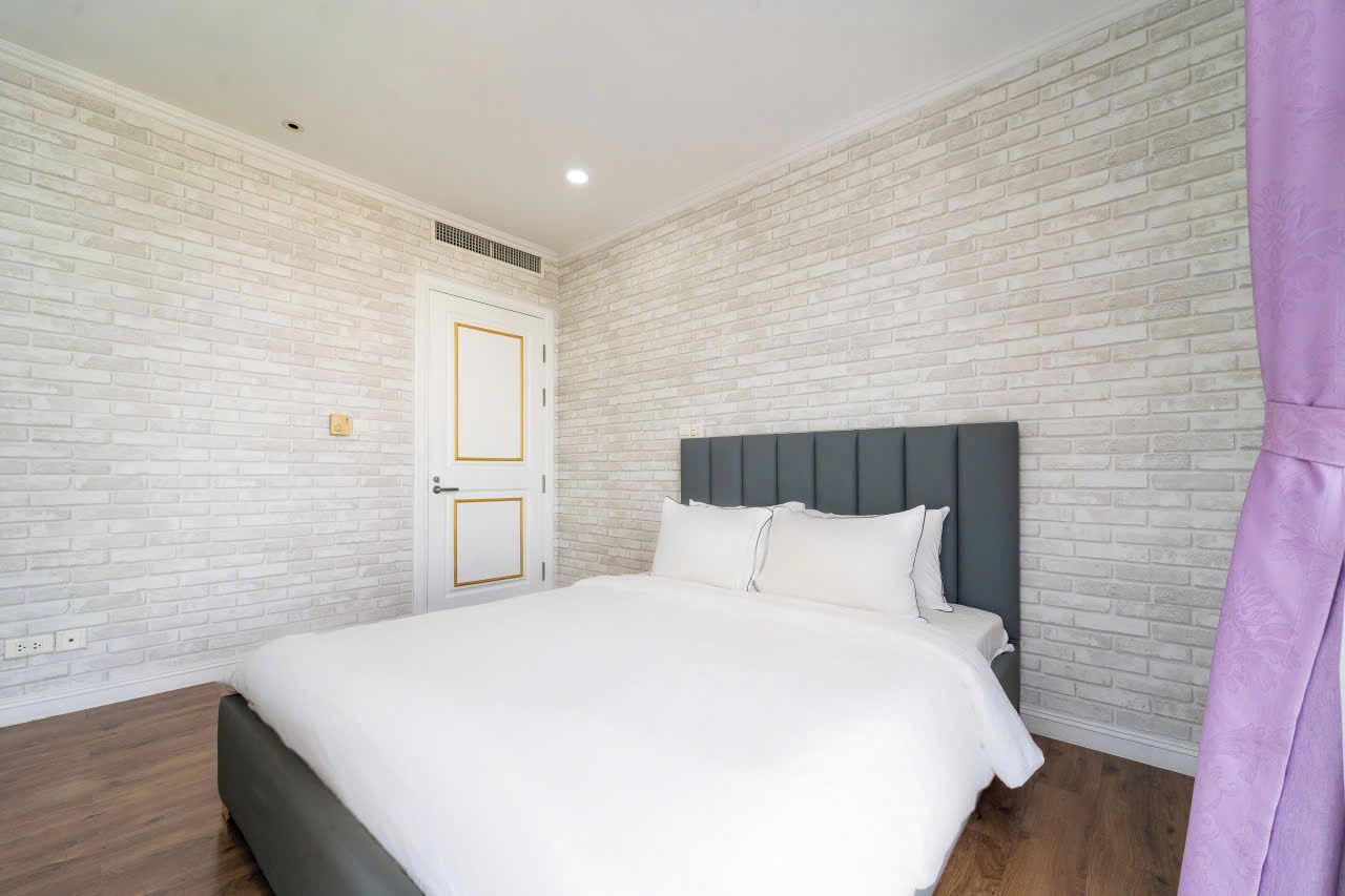 Un dormitorio con pared de ladrillo blanco, cabecero gris y colcha blanca.