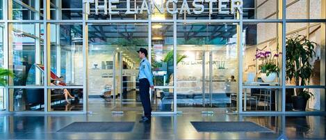 The entrance of The Langaster hotel with a modern glass facade, a security guard at the entrance, and potted plants inside.