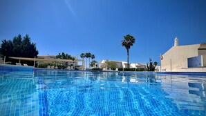 Ein klares, blaues Schwimmbecken mit gefliesten Rändern, umgeben von Liegestühlen und Palmen, unter blauem Himmel.
