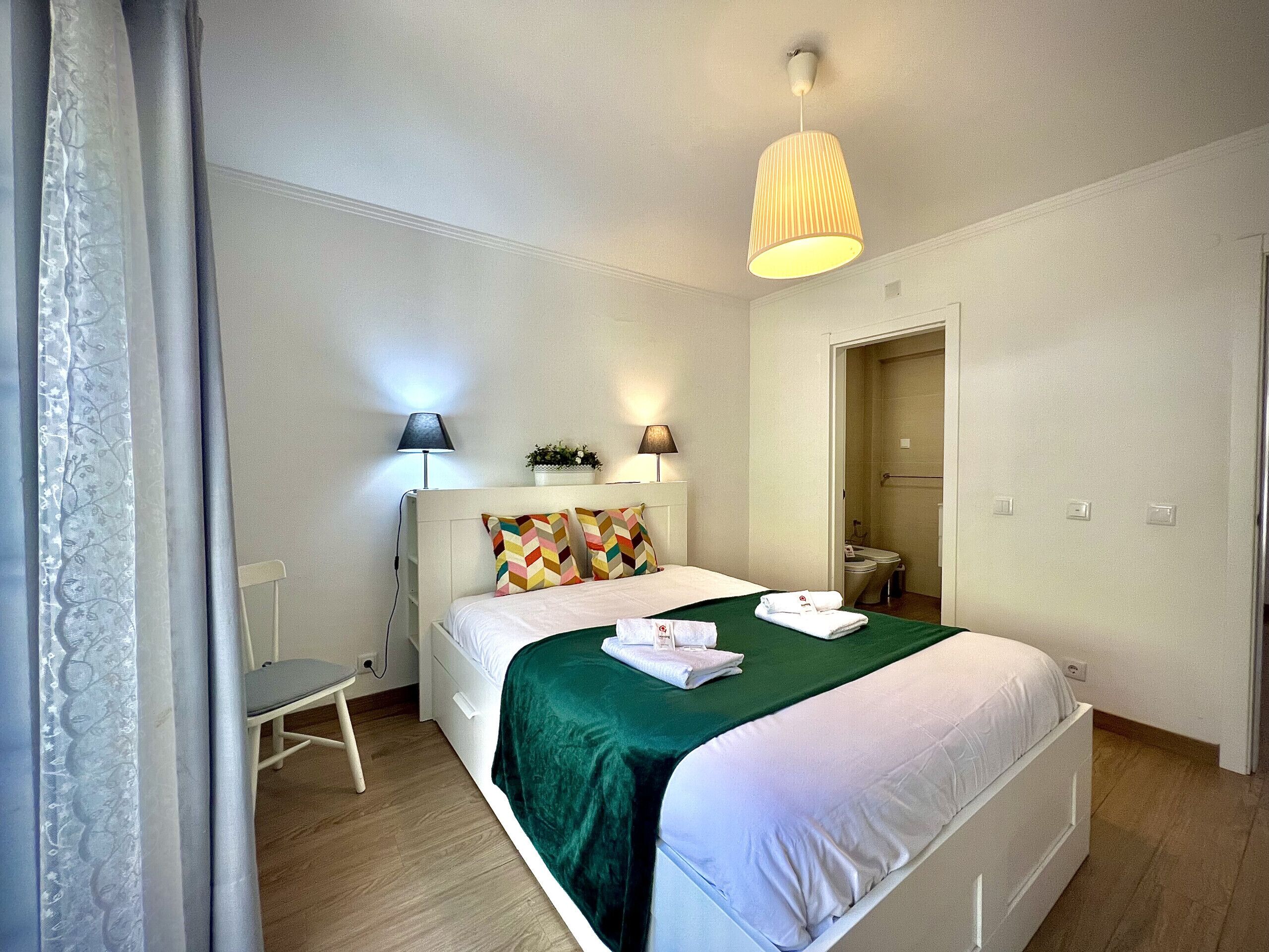 Una habitación de hotel con una cama, dos lámparas de noche, una silla y un baño visible a través de una puerta abierta.