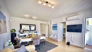 Ein modernes Wohnzimmer mit Sofa, Esstisch und Stühlen. Ein Fernseher ist an der Wand befestigt, und durch eine offene Tür ist eine Küchenzeile erkennbar.