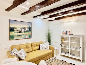 Una sala de estar con un sofá, un cuadro, un aparador con vajilla y vigas de madera a la vista.