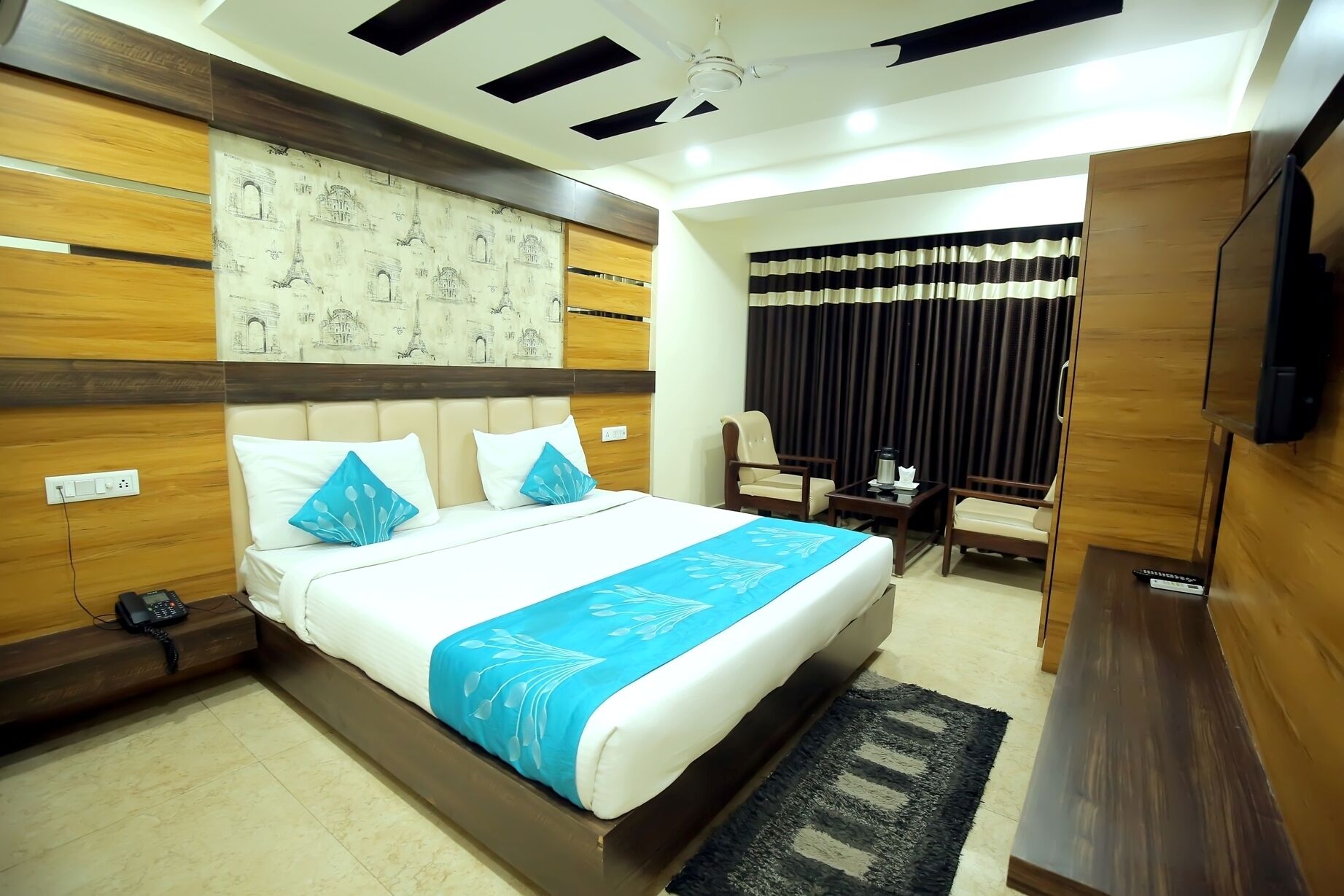Quarto de hotel com cama, poltrona, escrivaninha, televisão e janela com cortinas.