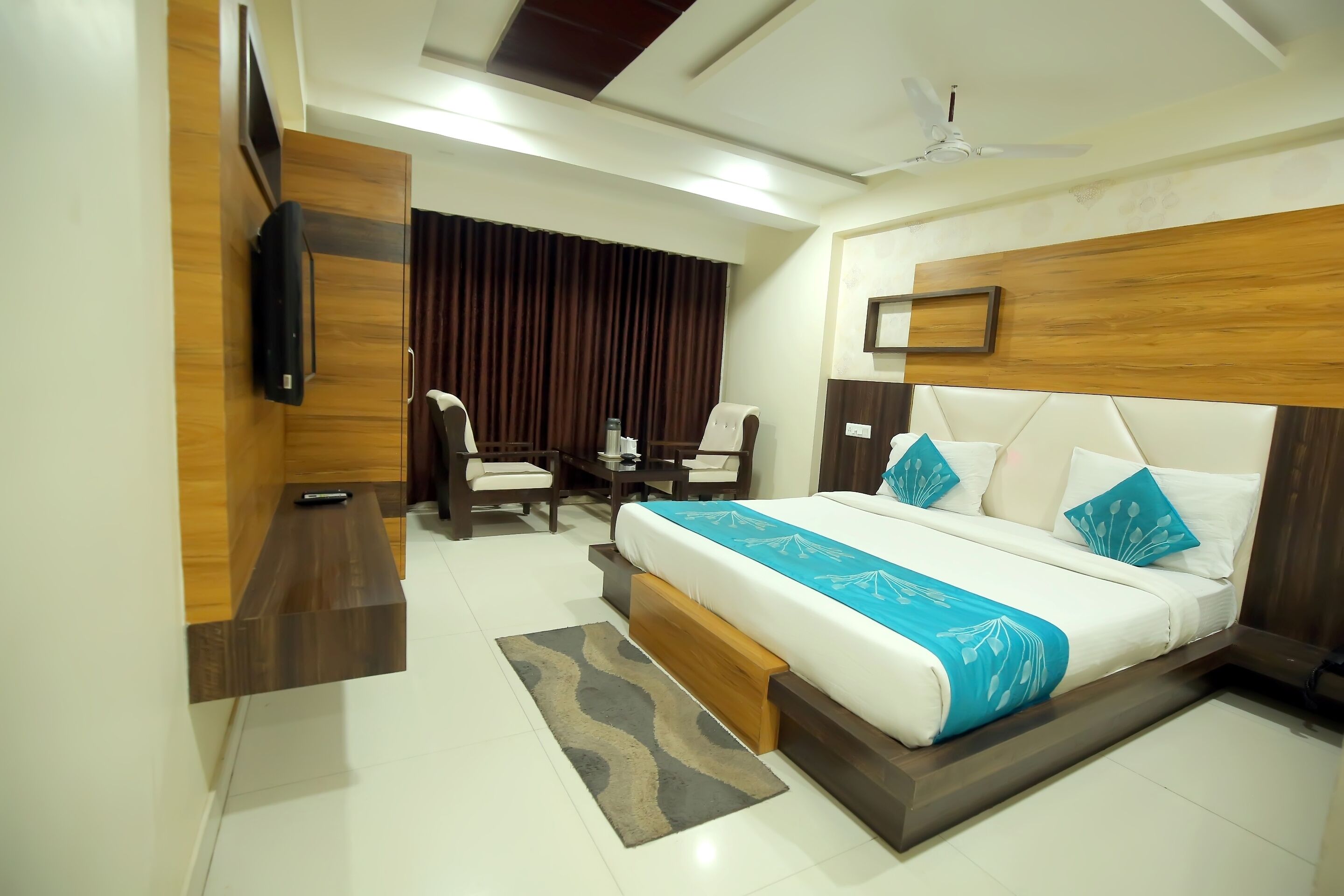A hotel room with a large bed, a desk, a chair, a TV mounted on the wall, and a ceiling fan.