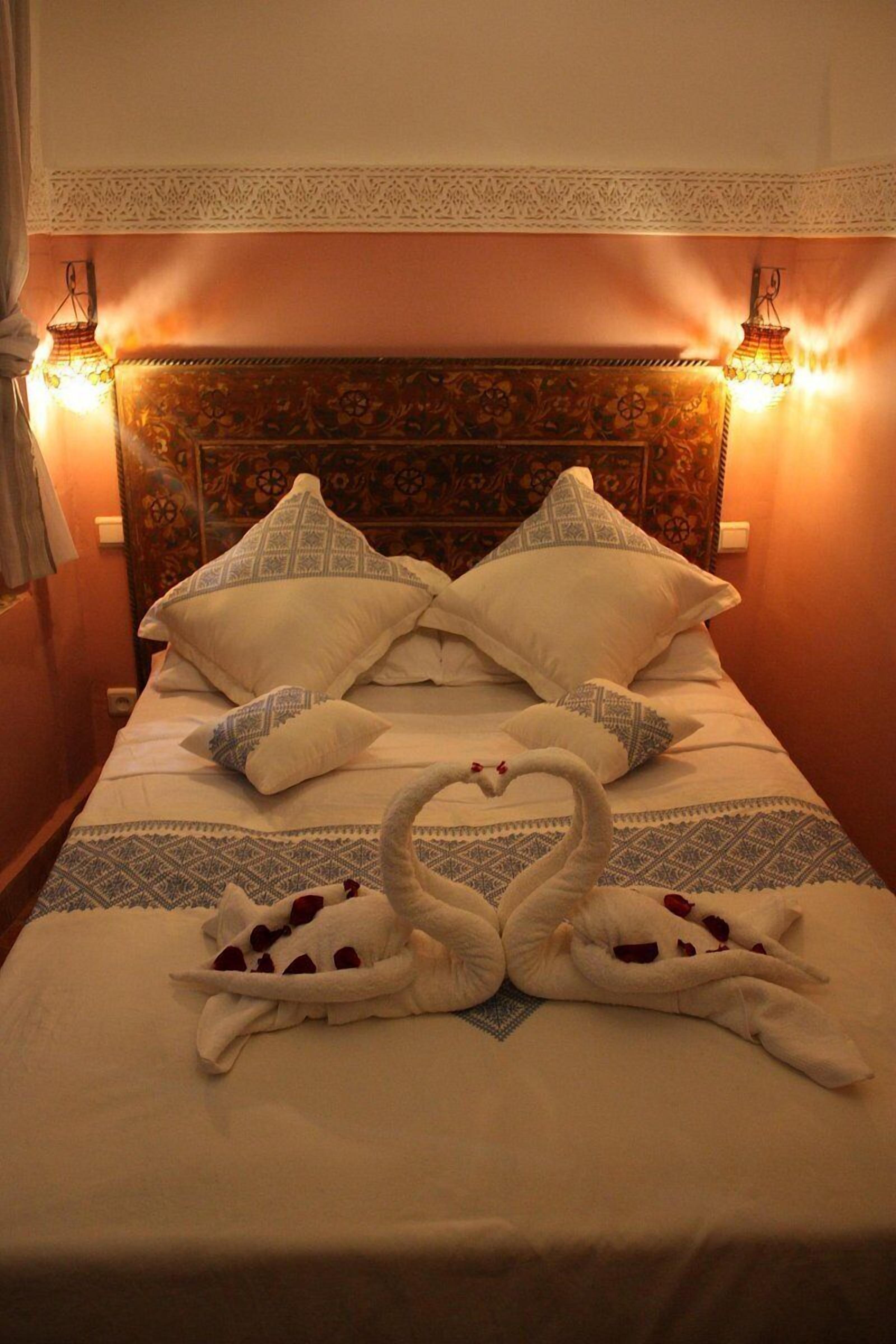 A bed with a decorative headboard, two wall-mounted lamps, and a swan-shaped towel arrangement.