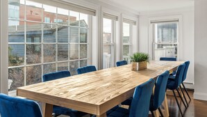 Une longue table en bois, des chaises bleues, de grandes fenêtres et une plante en pot.