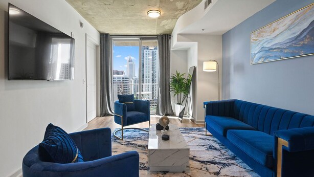 A modern living room with a large TV, two blue sofas, a coffee table, and a view of the city through large windows. - Royal Retreat (Atlanta)