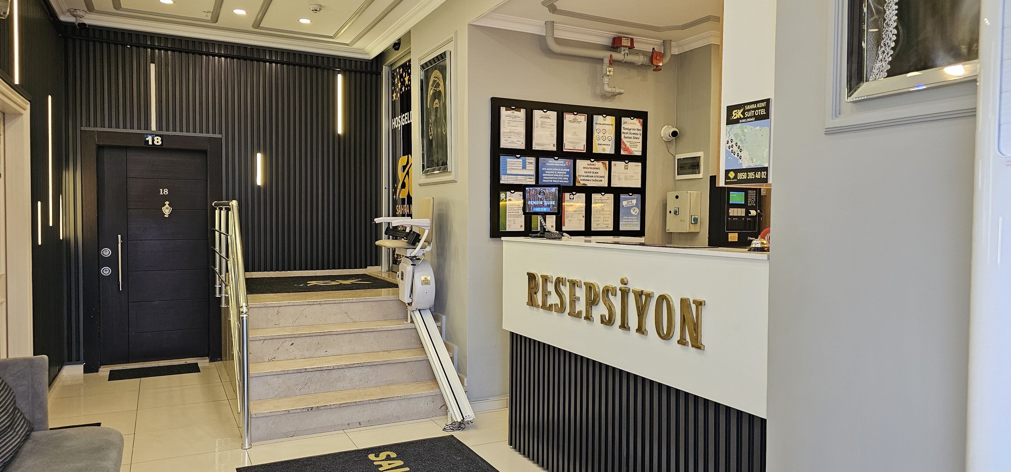 Zona de recepción de hotel con una escalera que sube al segundo piso, un mostrador de recepción y una pared con diversos certificados y avisos.
