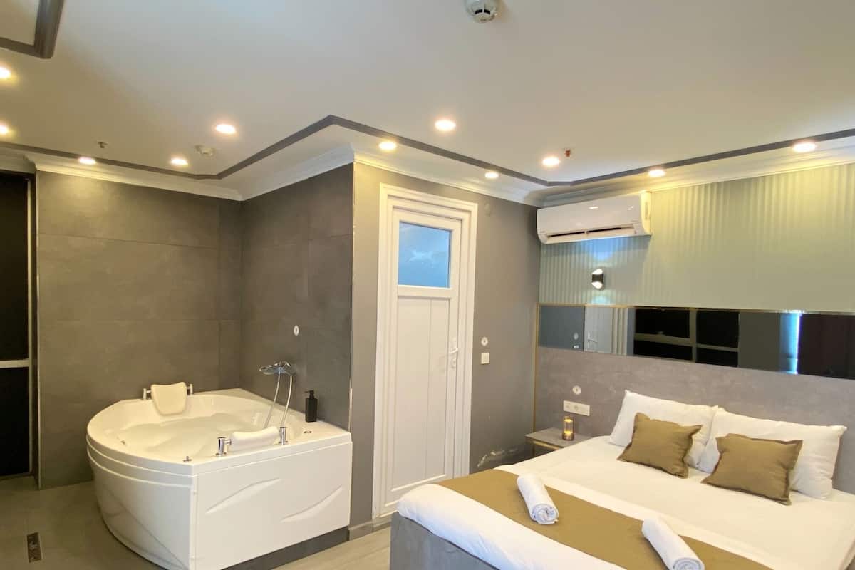 A modern hotel room with a large bathtub, a bed with a brown headboard, and a bedside table with a lamp.